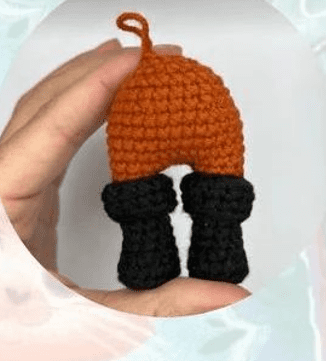 Una Mano Sostiene Una Pequeña Figura Tejida a Crochet De Un Par De Pantalones Rojos Con Botas Negras Que Se Asemeja Al Icónico Gato Con Botas De Shrek Amigurumi Los Pantalones Son Rojos Con Un Tinte Naranja Y Tienen Un Pequeño Lazo En La Parte Superior Las Botas Son Negras Y Parecen Estar Tejidas Por Separado Y Unidas a Los Pantalones