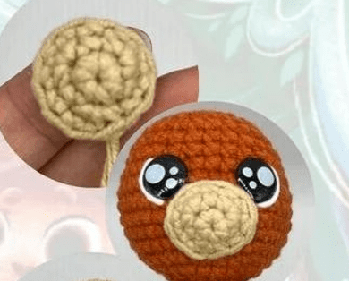 Una Imagen De Primer Plano Muestra Un Juguete Tejido a Crochet En Proceso El Amigurumi En Español Tiene Una Cara Naranja Y Ojos Grandes Y Redondos Con Reflejos Blancos En Una Mano Sostiene Una Pequeña Pieza De Ganchillo Separada Para El Juguete Que Es De Color Beige Más Claro El Fondo Está Borroso