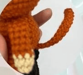 Primer Plano De Una Mano Que Sostiene Una Pequeña Figura De Un Animal Naranja Tejida a Crochet Posiblemente Un Zorro Con Detalles Blancos En La Punta De La Cola El Fondo Es Borroso Y Claro Llamando La Atención Sobre Lo Que Podría Ser Un Amigurumi De Gato Con Botas Inspirado En Shrek