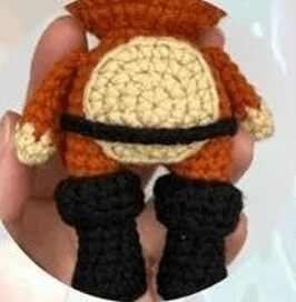 Una Persona Sosteniendo Un Muñeco Amigurumi En Español Tejido a Crochet Desde Atrás La Muñeca Tiene Un Cuerpo Naranja Con Un Parche Circular Beige En La Espalda Botas Negras Y Un Cinturón Negro La Figura Parece Estar Hecha a Mano Y Le Falta Una Cabeza