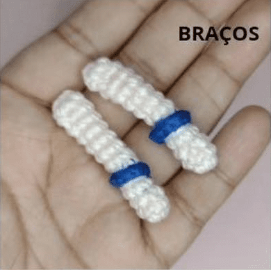 Primer Plano De Una Mano Que Sostiene Dos Pequeños Objetos Blancos De Crochet Con Anillos Azules Etiquetados Como braços En Portugués Que Significa brazos El Fondo Es Ligeramente Borroso Y De Color Violeta Claro Perfecto Para Cualquier Proyecto De Amigurumi Goku O Tutorial De Amigurumi Español