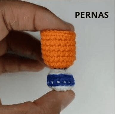 Una Mano Sostiene Pequeñas Piezas De Crochet Diseñadas Para Parecerse a Piernas La Parte Superior Es Naranja Y La Parte Inferior Es Azul Con Detalles En Blanco Que Recuerda a Un Amigurumi De Goku La Palabra pernas en Portugués Significa piernas Está Impresa En La Imagen