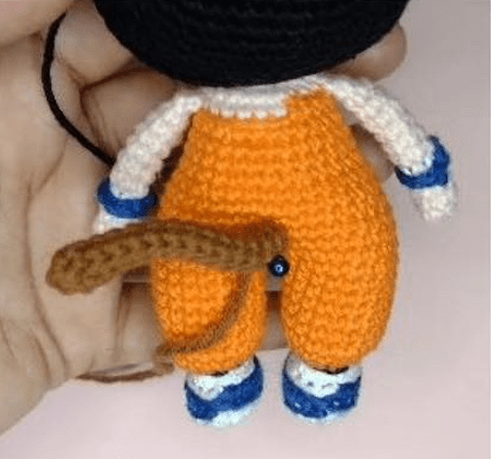 Un Pequeño Muñeco Amigurumi De Dragon Ball Con Un Mono Naranja Camisa Blanca Y Zapatos Azules El Muñeco Tiene Una Cola Marrón Unida a La Parte Posterior De Su Mono Que Recuerda Al Amigurumi Goku Lo Sostiene La Mano De Alguien Sobre Un Fondo Rosa Claro