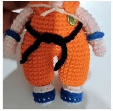 Una Muñeca De Crochet Vestida Con Un Conjunto Naranja Con Un Cinturón Negro Este Amigurumi Goku También Tiene Muñequeras Y Zapatos Azules Que Recuerdan a Dragon Ball La Parte Superior De La Muñeca No Se Ve En La Imagen El Fondo Es Completamente Blanco