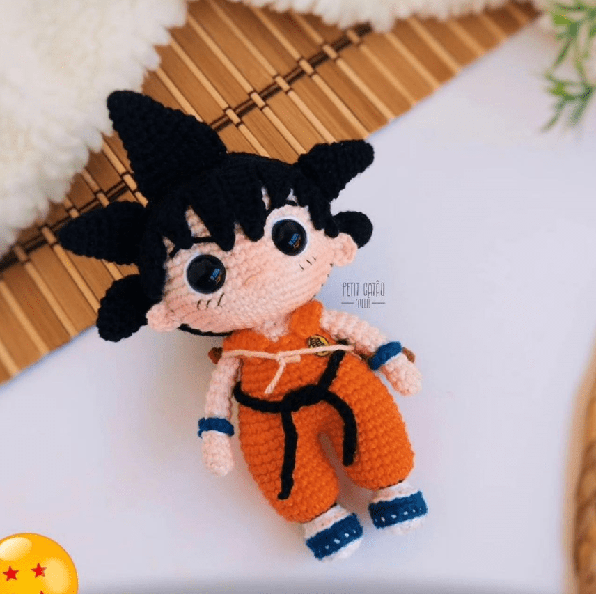 Un Muñeco Amigurumi Goku Que Se Asemeja a Un Personaje Animado Con Cabello Negro Puntiagudo Y Que Viste Un Traje Naranja De Artes Marciales Yace Sobre Una Estera De Bambú La Muñeca Tiene Ojos Grandes Y Expresivos Y Está Detallada Con Un Cinturón Y Zapatos Azules En La Esquina Aparece Una Pequeña Bola De Dos Estrellas