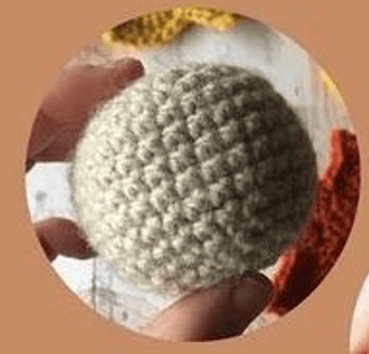 Un Primer Plano De Una Persona Sosteniendo Un Objeto Esférico Tejido a Mano Con Hilo De Color Claro Las Puntadas Texturizadas Son Claramente Visibles En El Fondo Borroso Hay Indicios De Otras Piezas De Crochet En Diferentes Colores Posiblemente Creaciones De Amigurumi Paso a Paso