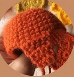 Imagen De Primer Plano De Una Persona Sosteniendo Una Acogedora Taza Tejida O De Ganchillo De Color Naranja Que Cubre Parcialmente Sus Dedos Al Fondo Se Pueden Ver Objetos Borrosos De Color Amarillo Y Naranja Que Se Asemejan a Vibrantes Creaciones De Amigurumi Paso a Paso