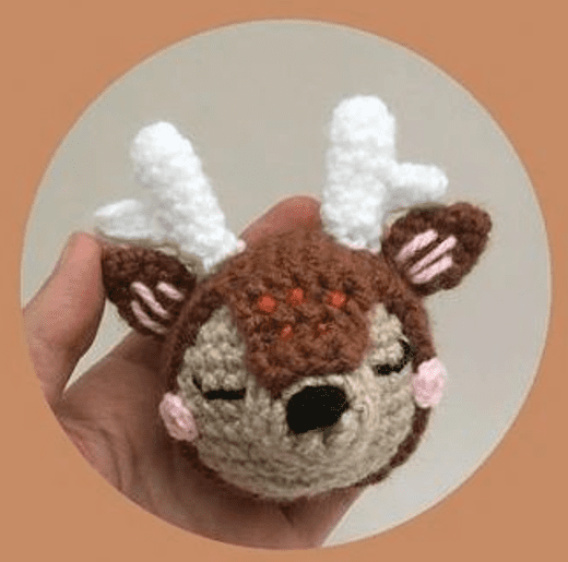 Una Mano Que Sostiene Una Pequeña Cabeza De Ciervo Amigurumi Hecha a Mano Con Astas Blancas Orejas Grandes Y Ojos Cerrados Sobre Un Fondo Beige El Ciervo Tiene Las Mejillas Rosadas Y La Nariz Negra Con Algunas Manchas Anaranjadas En La Frente