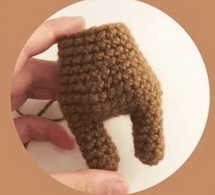 Primer Plano De Una Mano Que Sostiene Un Objeto De Crochet Parcialmente Terminado Que Se Asemeja a Las Patas De Un Pequeño Animal De Peluche O Muñeca Hecho Con Hilo Grueso De Color Marrón Esta Creación De Amigurumi Ciervo Se Presenta Sobre Un Fondo Blanquecino Sólido Dentro De Un Marco Circular