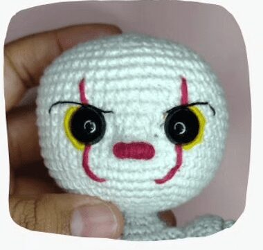 Una Imagen De Primer Plano De Una Mano Que Sostiene Una Cabeza De Muñeco De Ganchillo Que Parece Un Payaso La Muñeca Tiene Una Cara Blanca Con Grandes Ojos Negros Y Amarillos Líneas Rojas Que Van Desde Los Ojos Hasta La Boca Y Una Nariz Roja El Fondo Está Ligeramente Borroso