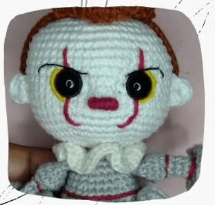 Un Muñeco De Ganchillo Que Se Asemeja a Un Payaso Con Distintivos Ojos Rojos Y Amarillos Pintura Facial Blanca Y Marcas Rojas La Muñeca Tiene Cabello Castaño Un Cuello Blanco Con Volantes Y Un Traje Gris Con Detalles En Rojo Se Ve Parcialmente La Mano De Una Persona Que Sostiene La Muñeca