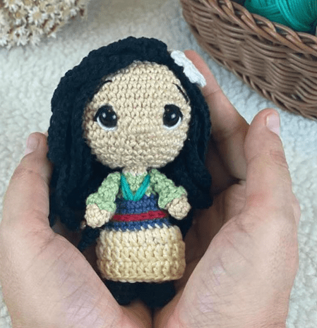 Una Pequeña Muñeca Tejida a Crochet Con Pelo Largo Y Negro Ojos Grandes Y Negros Y Una Flor Blanca En La Cabeza Se Sostiene Suavemente Entre Dos Manos La Muñeca Está Vestida Con Un Traje Verde Y Azul Con Una Cinturilla Roja En El Fondo Se Ve Una Cesta De Mimbre Con Hilo