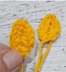 Dos Pequeñas Formas De Hojas Amarillas Tejidas a Crochet Cada Una Con Una Cola De Hilo Colocadas Sobre Una Superficie Blanca Con Textura Una Mano Con El Pulgar Visible Sostiene El Borde De Una De Las Piezas Tejidas a Crochet