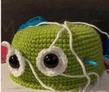 🩷 Cómo Tejer un Amigurumi de Alíen de Toy Story: Patrón Gratis en español 👽 - Un proyecto de crochet verde parcialmente terminado con ojos negros grandes y redondos colocados en círculos blancos. Se enredan hebras sueltas de hilo blanco a su alrededor y se coloca un ganchillo azul. El fondo está difuminado y se ve un poco de hilo azul. Kit de materiales básicos para tejer amigurumis sobre una mesa de madera, incluyendo ovillos de colores, ganchillos, ojos de seguridad y relleno.