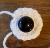 🩷 Cómo Tejer un Amigurumi de Alíen de Toy Story: Patrón Gratis en español 👽 - Sobre una superficie de madera se coloca una pequeña flor blanca, redonda y tejida a crochet, con un gran botón negro en el centro. De la flor se extiende una hebra de hilo blanco.