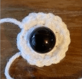 Sobre Una Superficie De Madera Se Coloca Una Pequeña Flor Blanca Redonda Y Tejida a Crochet Con Un Gran Botón Negro En El Centro De La Flor Se Extiende Una Hebra De Hilo Blanco