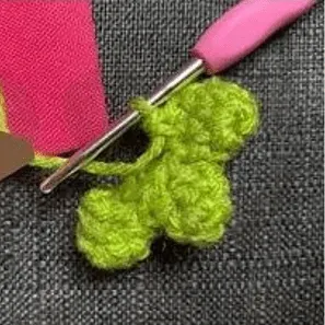🩷 Cómo Tejer un Amigurumi de Alíen de Toy Story: Patrón Gratis en español 👽 - Primer plano de un proyecto de crochet, en el que se trabaja con hilo verde formando una flor. Se inserta un ganchillo con mango rosa en los puntos. El fondo es una tela gris texturizada. Fila diagonal de ganchillos de colores con mangos ergonómicos, rodeados de ovillos de lana y marcadores de puntos sobre fondo blanco.