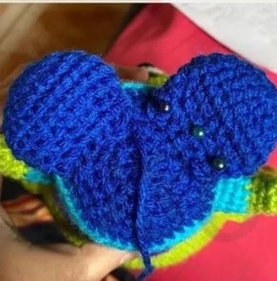 🩷 Cómo Tejer un Amigurumi de Alíen de Toy Story: Patrón Gratis en español 👽 - Primer plano de una persona que sostiene un proyecto de crochet en proceso, con hilo azul con detalles verdes y amarillos. El proyecto parece implicar la creación de un objeto de peluche, posiblemente un animal, con alfileres colocados en la sección azul para su ensamblaje. Composición plana de ovillos de lana en tonos púrpuras y lilas, decorada con flores de lavanda y corazones.