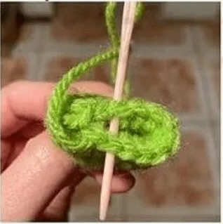 🩷 Cómo Tejer un Amigurumi de Alíen de Toy Story: Patrón Gratis en español 👽 - Imagen en primer plano de una persona que sostiene un palo de madera o una aguja de tejer verticalmente a través del centro de un bucle de hilo verde brillante, lo que posiblemente indica un paso en un proceso de tejido o crochet. El fondo muestra un piso de baldosas. Varias tijeras de bordado y ovillos de lana pastel sobre un fondo blanco con marcadores de punto y botones de colores.