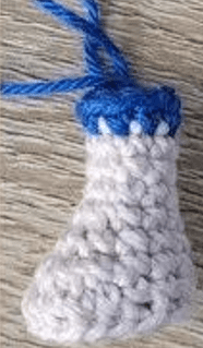 Un Pequeño Artículo Tejido a Crochet Con Forma De Media O Calcetín Blanco Con Un Dobladillo Azul En La Parte Superior En La Parte Superior Se Encuentra Un Lazo De Hilo Azul Para Colgarlo Se Encuentra Apoyado Sobre Una Superficie De Madera