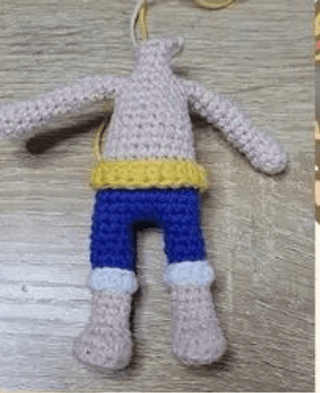 Figura Tejida a Mano Con El Torso Sin Cabeza Brazos Extendidos Y Pantalones Azules Con Puños Blancos Con Un Cinturón Amarillo La Figura Yace Sobre Una Superficie De Madera Parece Estar En Proceso De Fabricación