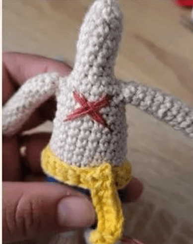 Primer Plano De Un Patricio Estrella De Bob Esponja Tejido a Crochet El Juguete Es De Color Beige Con Una X Roja En El Pecho Y Lleva Un Cinturón Amarillo La Mano De Una Persona Lo Sostiene El Fondo Es Una Superficie De Madera De Color Claro