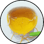 Un Artículo Amarillo Parcialmente Tejido a Crochet Con Ribete Blanco Y Un Ganchillo Con Mango Verde Adjunto El Proyecto Está En Marcha Con Varios Hilos De Hilo Visibles La Imagen Está Encerrada En Un Marco Circular