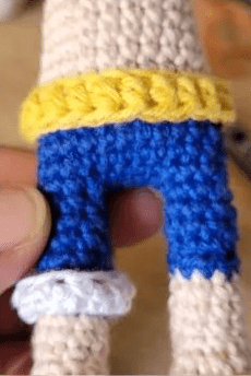 Primer Plano Del Torso Y Las Piernas De Una Muñeca Tejida a Crochet La Muñeca Lleva Pantalones Azules Con Un Cinturón Amarillo La Pierna Derecha Tiene Un Puño Azul Claro En La Parte Inferior Se Ven Parcialmente El Pulgar Y El índice De Una Persona Que Sostiene La Muñeca