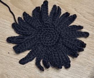 Se Muestra Una Flor Tejida a Crochet Negra Con Un Centro En Espiral Y Pétalos Alargados Sobre Una Superficie De Madera Un Hilo De Lana Suelto Se Extiende Desde El Pétalo Superior Izquierdo