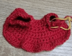 Sobre Una Superficie De Madera Se Encuentra Un Pequeño Trozo De Tela Tejida a Crochet De Color Rojo Con Un Borde Fruncido Una Parte De La Tela Tiene Un Hilo Amarillo Enhebrado Posiblemente Para Marcar Puntos O Asegurar La Pieza