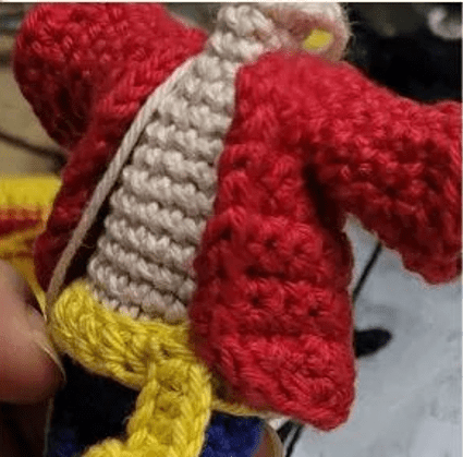 Figura Tejida a Crochet Con Chaqueta Roja Camisa O Torso Beige Cinturón Amarillo Y Pantalón Azul La Figura Parece Hecha a Mano Con Una Mezcla De Puntos De Crochet Y Sostenida En La Mano De Una Persona