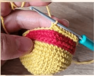 Primer Plano De Una Mano Que Sostiene Un Proyecto De Crochet Sin Terminar Con Un Ganchillo El Hilo Es Amarillo Con Una Raya Roja El Fondo Es Una Superficie De Madera