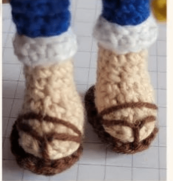 Primer Plano De Los Pies De Una Muñeca Amigurumi Con Sandalias Tejidas a Crochet Los Pies Son De Color Beige Claro Y Las Sandalias Son Marrones Con Detalles En Blanco Y Azul Visibles En La Parte Superior De La Imagen Posiblemente Parte De La Ropa De La Muñeca La Superficie Debajo Tiene Un Patrón De Cuadrícula