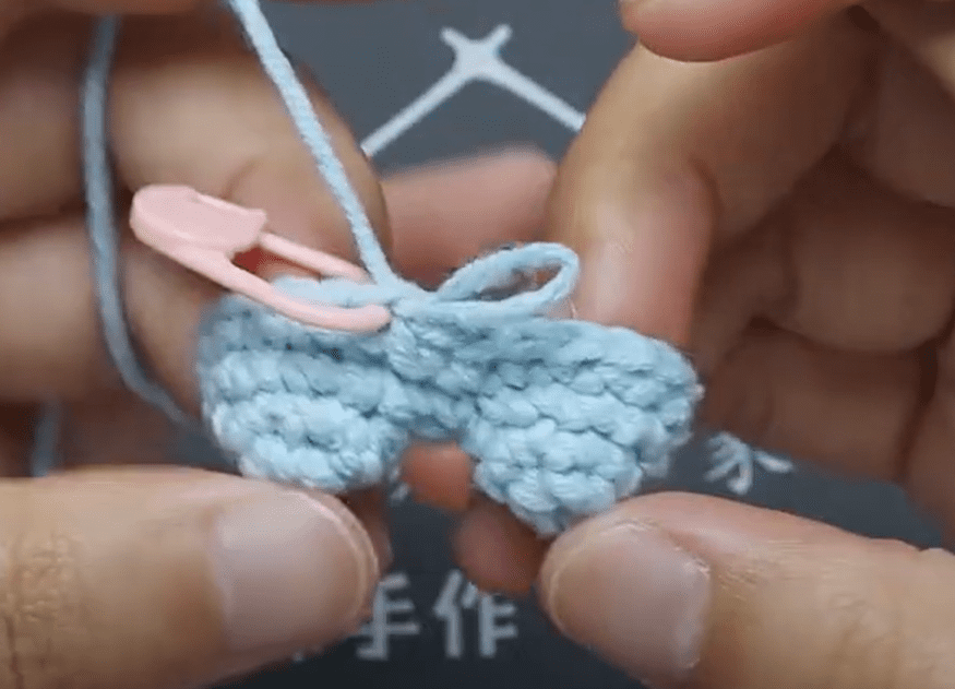 Primer Plano De Unas Manos Que Sostienen Una Pieza De Crochet Hecha Con Hilo Azul Claro Hay Un Marcador De Punto Rosa Adherido Al Crochet Y El Fondo Muestra Una Pizarra Borrosa Con Marcas Blancas La Pieza Está En Proceso Y El Hilo Se Enhebra a Través Del Ganchillo