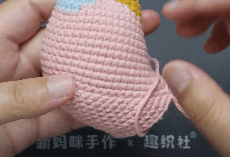 Primer Plano De Unas Manos Tejiendo a Crochet Un Trozo De Hilo Rosa El Crochet Crea Una Forma Cilíndrica Con Textura El Fondo Es Gris Con Algunas Marcas Blancas Que Quedan Parcialmente Ocultas Por Las Manos