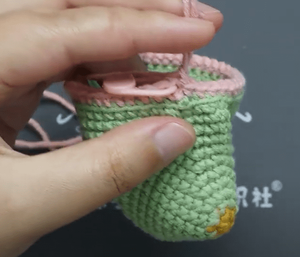 Primer Plano De Una Mano Que Sostiene Un Pequeño Proyecto De Crochet Parcialmente Terminado Con Hilo Verde Y Rosa La Pieza De Crochet Parece Ser Cilíndrica Con Algunos Puntos En La Parte Inferior Y Un Bucle En La Parte Superior