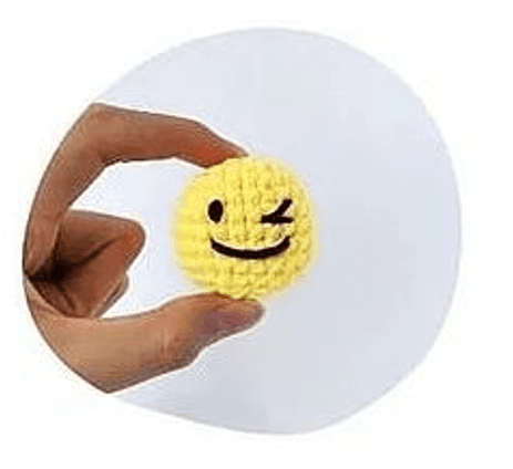 Una Mano Sostiene Una Pequeña Pelota Redonda De Color Amarillo Tejida Con Una Cara Que Guiña El Ojo La Pelota Tiene Un Ojo Abierto Otro Cerrado Y Una Sonrisa