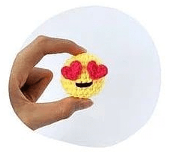 Una Mano Que Sostiene Un Objeto Redondo Tejido a Crochet De Color Amarillo Con Una Cara Sonriente Y Ojos En Forma De Corazón Similar Al Emoji De ojos De Corazón Sobre Un Fondo Blanco