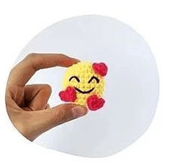 Una Mano Sostiene Un Objeto Pequeño Redondo Y Tejido a Crochet De Color Amarillo Con Una Carita Sonriente La Carita Sonriente Tiene Los Ojos Cerrados Y Está Adornada Con Tres Corazones Rojos El Fondo Es Blanco Liso Lo Que Enfatiza La Naturaleza Tierna Y Artesanal Del Artículo Tejido a Crochet