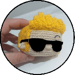 Una Mano Que Sostiene Una Pequeña Figura Tejida a Crochet Con Cabello Amarillo Brillante Gafas De Sol Y Una Cara Beige La Figura Parece Llevar Una Máscara Negra Sobre Los Ojos El Fondo Es Liso Y De Colores Claros