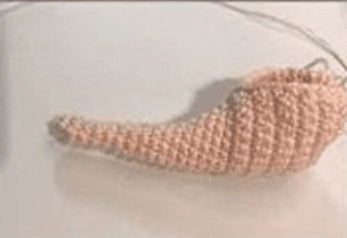 Un Objeto Parcialmente Tejido a Crochet Que Se Asemeja a La Forma De Una Zanahoria O a Un Sombrero Puntiagudo Caprichoso Yace Sobre Una Superficie Plana Elaborado Con Un Hilo De Color Melocotón Un Ganchillo De Metal Está Unido Al Trabajo En Curso