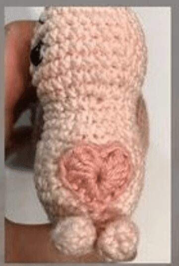 Una Mano Sostiene Un Pequeño Animal De Crochet Rosa Posiblemente Un Cerdo Que Se Muestra Desde Atrás El Juguete Presenta Una Nariz Rosa Circular Con Un Detalle De Corazón La Textura Y Los Detalles Del Crochet Son Claramente Visibles Fondo Blanco