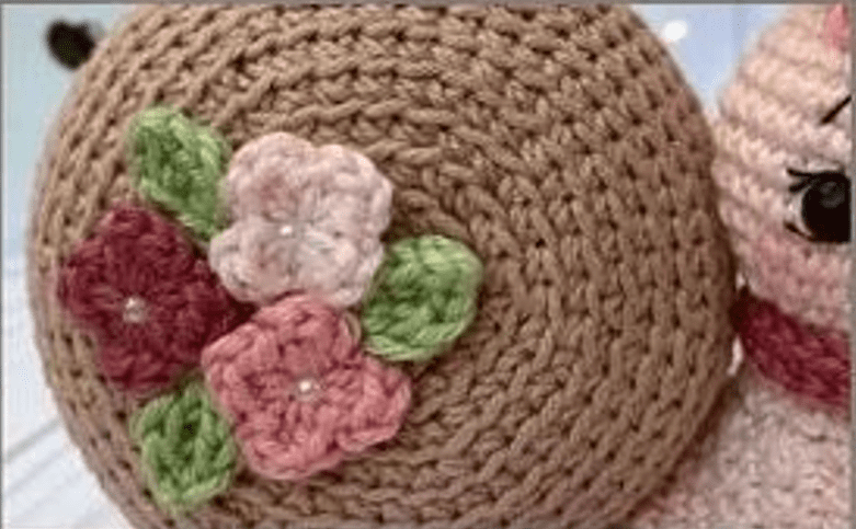 Primer Plano De Un Gorro De Muñeca Tejido a Crochet Con Intrincadas Puntadas El Gorro Está Adornado Con Tres Pequeñas Flores Tejidas a Crochet En Rosa Y Burdeos Con Hojas Verdes Que Crean Un Detalle Encantador Algunos De Los Rasgos Faciales De La Muñeca Son Parcialmente Visibles En El Lado Derecho De La Imagen