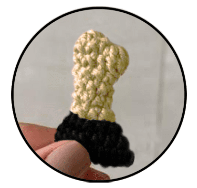 Una Mano Sostiene Un Pequeño Artículo Tejido a Crochet Que Parece Un Trofeo O Un Premio La Parte Superior Está Tejida a Crochet Con Hilo Amarillo Y La Base Está Construida Con Hilo Negro El Artículo Está Dentro De Un Marco Circular Sobre Un Fondo Liso Y De Color Claro
