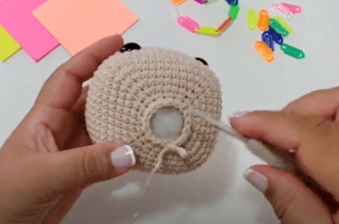 Una Persona Sostiene Un Objeto Tejido a Crochet Con Una Pequeña Abertura Que Parece Ser Un Peluche Y Trabaja En él Con Un Ganchillo Sobre La Mesa Del Fondo Hay Clips De Papel Y Notas Adhesivas De Colores