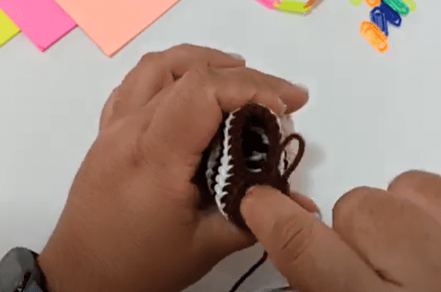 Se Ven Dos Manos Tejiendo O Haciendo Crochet Un Objeto Pequeño Con Hilo Marrón Y Blanco En El Fondo Sobre Una Superficie Blanca Hay Papeles Y Clips De Colores El Dedo índice Izquierdo De La Persona Está Dentro Del Objeto Guiando El Proceso