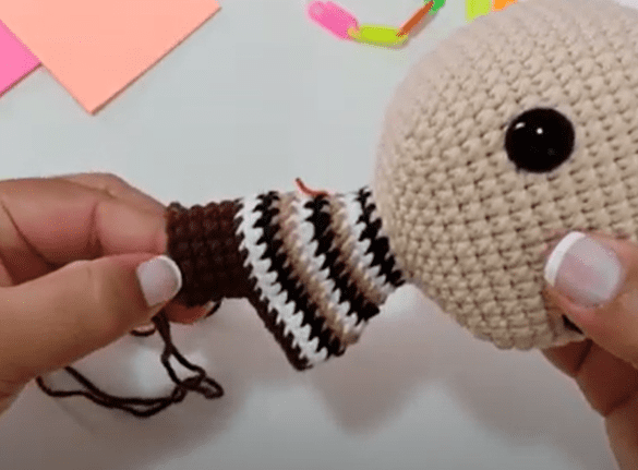 Una Persona Sostiene Un Muñeco Amigurumi Parcialmente Tejido a Crochet El Muñeco Tiene Una Cabeza Beige Con Un Gran Ojo Negro Y Está Trabajando En El Cuerpo Con Rayas Marrones Blancas Y Negras En El Fondo Hay Clips De Papel De Colores Y Notas Adhesivas