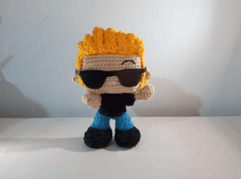 Una Muñeca De Ganchillo Con Pelo Amarillo Puntiagudo Y Gafas De Sol Negras La Muñeca Lleva Una Camisa Negra Pantalones Azules Y Zapatos Negros Se Encuentra Sobre Una Superficie Blanca Sobre Un Fondo Liso De Color Claro