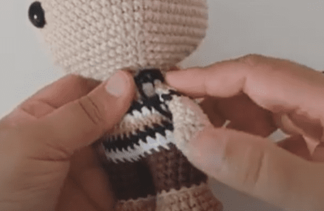 Un Par De Manos Sostienen Una Muñeca Tejida a Crochet a La Que Le Colocan Un Pequeño Objeto Negro En La Espalda La Muñeca Parece Hecha a Mano Y Presenta Un Patrón De Rayas En Su Cuerpo Con Tonos De Marrón Beige Y Negro El Fondo Es Liso Y Está Desenfocado