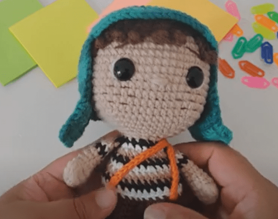 Una Muñeca Tejida a Crochet Con Un Sombrero Verde Y Una Camisa a Rayas Se Sostiene Suavemente En Las Manos De Alguien En El Fondo Cuadrados De Papel De Colores Y Clips De Papel Multicolores Están Dispersos Sobre Una Superficie Blanca